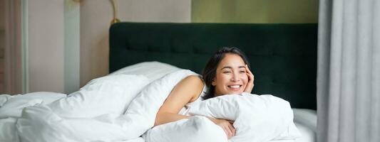 Happy mornings. Beautiful korean woman lying in bed, smiling, waking up upbeat, cover herself with warm duvet photo