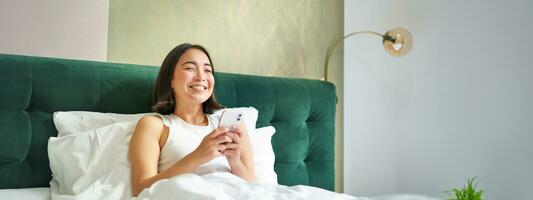 Beautiful smiling asian woman in bed, reading message on smartphone and laughing, enjoying lazy day in bedroom, using mobile phone photo
