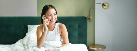sonriente coreano niña en cama, negociaciones en móvil teléfono, haciendo un teléfono llamar, perezoso Mañana como asiático mujer pedidos entrega vía teléfono inteligente foto