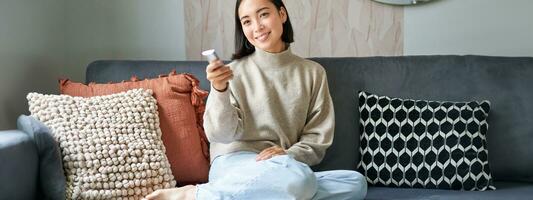 Beautiful asian girl in cozy home, watching television, holding remote and switching channels, sitting on sofa photo