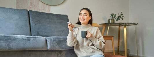 Portrait of smiling korean woman sitting near tv, holding remote and switching channels while drinking hot coffee photo