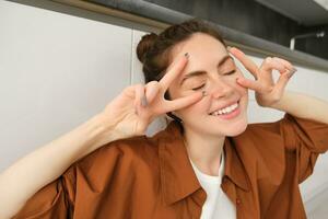 Close up portrait of beautiful young woman, enjoying her songs playlist in wireless headphones, listening to music and showing peace, v-sign gestures, smiling happily photo