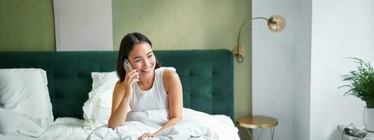 Smiling korean girl talks on mobile phone and lying in bed. Cute woman answers telephone call, holds smartphone, relaxing in her bed photo