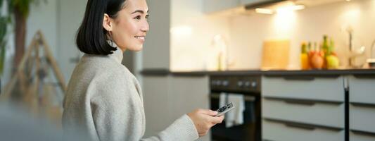 Close up portrait of smiling asian woman watching television, sitting on sofa with remote and switching chanel, looking relaxed photo