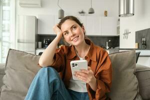 sonriente morena mujer sentado en sofá con teléfono inteligente, mirando considerado, pensando mientras utilizando móvil teléfono aplicación, ordenando en solicitud, haciendo en línea compras y descansando a hogar foto