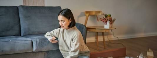 Vertical shot of young woman in cozy home working on laptop, using smartphone and drinking coffee, sitting on floor near sofa photo