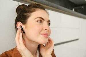 Close up photo of young smiling brunette woman, puts on her headphones, listens to music in wireless earphones, sits in the kitchen and enjoys sound quality