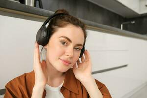 Close up portrait of beautiful brunette woman, listens to music in wireless headphones, has new earphones on her head, smiling and looking happy photo