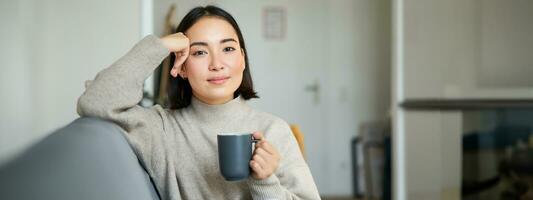 Smiling asian woman sitting on sofa with her mug, drinking coffee at home and relaxing after work, looking calm and cozy photo