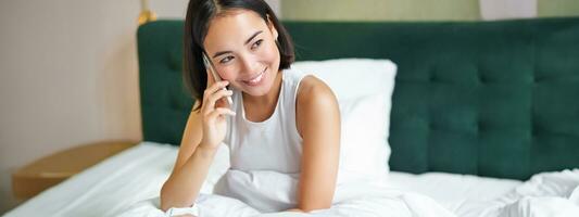 sonriente coreano niña negociaciones en móvil teléfono y acostado en cama. linda mujer respuestas teléfono llamar, sostiene teléfono inteligente, relajante en su cama foto