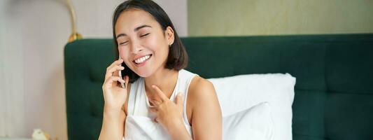 Close up portrait of cute asian girl in bed, talking on mobile phone with happy smiling face. Woman waking up and making a telephone call photo