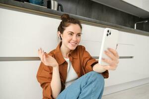 Friendly smiling woman talking on mobile phone app, using wireless earphones, looking at screen, online chatting, video calling friend, sitting on kitchen floor photo