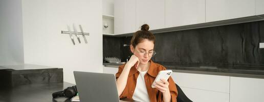 Portrait of young self-employed woman working on start up from home, freelancer programmer with computer, doing her task on computer, checking her mobile phone, reading message on smartphone photo