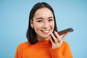 Portrait of asian woman records voice message, talks on speakerphone, holds mobile phone near mouth while speaking, translates her speech on app, blue background photo