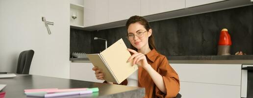 Portrait of young self-employed freelance tutor, sitting in kitchen at home, holding notebook with study notes, revising for university photo