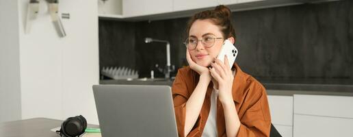 Portrait of young self-employed woman, entrepreneur working from home, freelancer calling client. Girl making an order, talking to someone on phone, sitting in kitchen with laptop photo