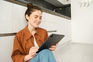 retrato de bonito joven mujer a hogar, sentado en cocina piso, mirando a digital tableta, utilizando artilugio, leyendo en dispositivo foto