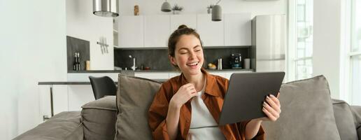 Portrait of woman laughing over funny video on digital tablet, watching movie on her gadget streaming service, sitting on sofa at home, reading e-book photo