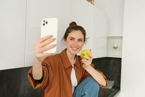 retrato de feliz, sonriente joven mujer registros sí misma, toma selfie mientras comiendo un manzana en el cocina, utilizando teléfono inteligente aplicación, hace fotos con móvil teléfono