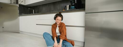 Portrait of young beautiful woman in wireless headphones, sitting on kitchen floor, vibing with favourite music in earphones photo