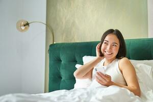 linda coreano niña en cama, participación teléfono inteligente, sensación contento y complacido, gasto Mañana en cama, disfrutando surf red en móvil teléfono foto