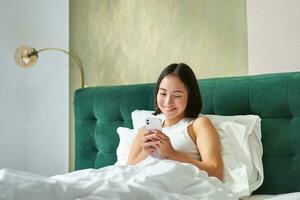 linda coreano niña en cama, participación teléfono inteligente, sensación contento y complacido, gasto Mañana en cama, disfrutando surf red en móvil teléfono foto