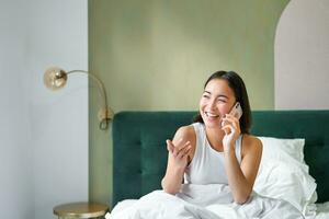 Beautiful smiling asian girl talking on mobile phone, lying in bed with pleased happy face, speaking to someone on telephone photo