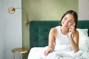 sonriente coreano niña en cama, negociaciones en móvil teléfono, haciendo un teléfono llamar, perezoso Mañana como asiático mujer pedidos entrega vía teléfono inteligente foto