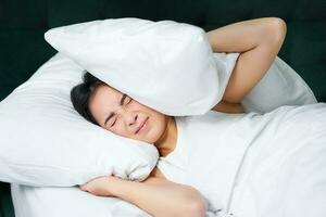 Sad girl with insomnia. Young asian woman lying in bed, cover her ears with pillow, cant sleep, partner snors, loud noise at night disturbing her photo