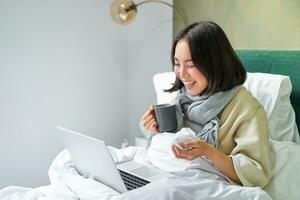 Portrait of asian sick girl, lying in bed with cold, having flu, talking to laptop, video chat, working from home on remote while on sick leave photo