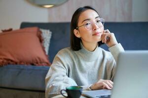 Trabajadores por cuenta propia joven sonriente mujer, persona de libre dedicación quedarse a hogar, trabajando en remoto desde computadora portátil, vistiendo anteojos, sentado en vivo habitación foto