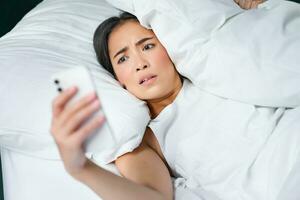 Portrait of asian woman waking up in bed, looking shocked at mobile phone, realise she overslept photo