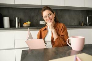 Portrait of beautiful young woman working from home, freelance tutor preparing for lesson. Student sitting in kitchen with folder, reading documents photo