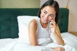 sonriente coreano niña negociaciones en móvil teléfono y acostado en cama. linda mujer respuestas teléfono llamar, sostiene teléfono inteligente, relajante en su cama foto