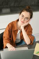 Vertical shot of beautiful woman working from home, student doing homework in kitchen, using laptop, looking at camera and smiling photo