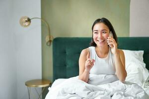 hermosa sonriente asiático niña hablando en móvil teléfono, acostado en cama con satisfecho contento rostro, Hablando a alguien en teléfono foto