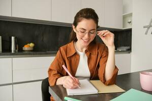 Portrait of young woman student, girl studying at home, working remote in kitchen, taking notes, sitting indoors photo