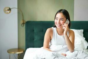 sonriente coreano niña en cama, negociaciones en móvil teléfono, haciendo un teléfono llamar, perezoso Mañana como asiático mujer pedidos entrega vía teléfono inteligente foto