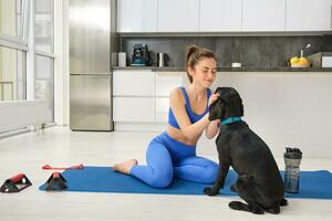 Image of young woman does workout from home, sits on yoga mat in living room and plays with her black dog, puppy distracts girl from doing exercises photo