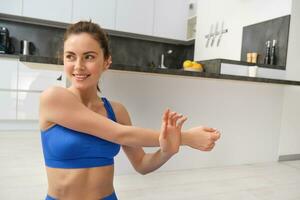 Close up shot of woman doing workout from home, warm-up stretching hands, does fitness exercises in blue sportsbra and smiling photo