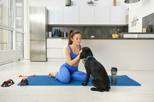 Image of young woman does workout from home, sits on yoga mat in living room and plays with her black dog, puppy distracts girl from doing exercises photo