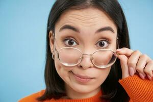 Close up portrait of amazed girl, looks closer at camera in glasses, standing against blue background photo