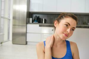 Close up portrait of young woman does fitness at home, touches her neck, rubs shoulders after workout training session photo