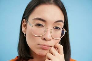 Close up portrait of asian woman looks intrigued, wears glasses, squints thoughtful, thinking, making assumption, standing over blue background photo