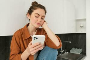 Image of modern stylish girl sits on kitchen counter with phone, looks at her mobile screen, checks messages, scrolling social media app on smartphone photo