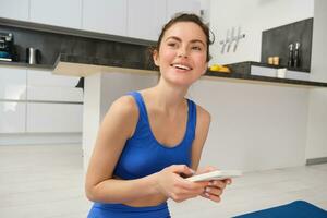 Portrait of healthy young sporswoman, smiling and holding smartphone, doing workout training at home in her house, fitness exercises photo