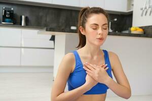 Image of calm and relaxed woman meditating, doing breathing practices, holding hands on chest during yoga session at home photo
