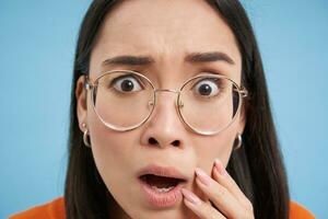 Close up portrait of japanese girl in glasses, looks shocked and concerned at camera, gasps worried, stands over blue background photo