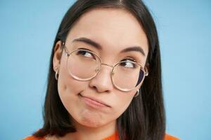 retrato de japonés mujer en anteojos, mira considerado, reflexiona, pensando con grave rostro, en pie terminado azul antecedentes foto