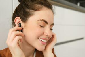 retrato de hermosa mujer disfruta escuchando a música o podcast en inalámbrico negro auriculares, utilizando auriculares, sonriente y mirando feliz, sentado a hogar en cocina piso foto